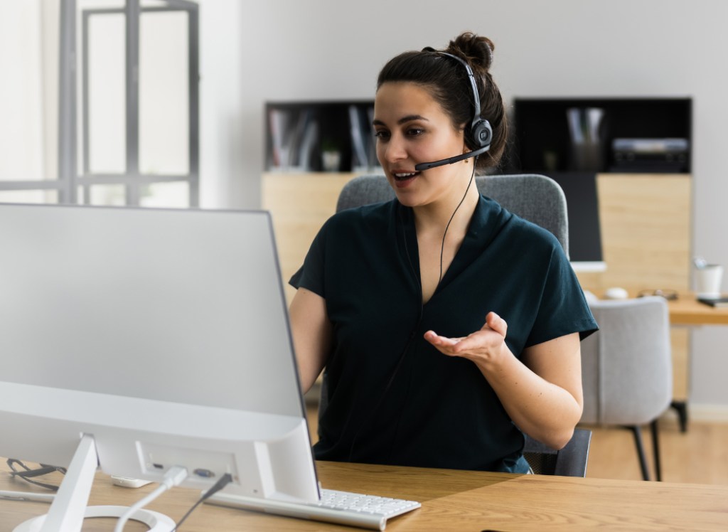 a woman talking into her headset sits at a table in front of a computer