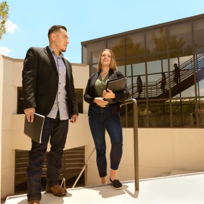 A man and a woman stand in front of a building holding laptops.