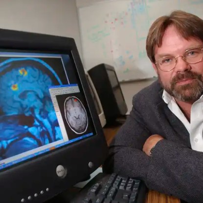 A psychology professor poses next to a computer displaying brain scans — NMSU Global Campus