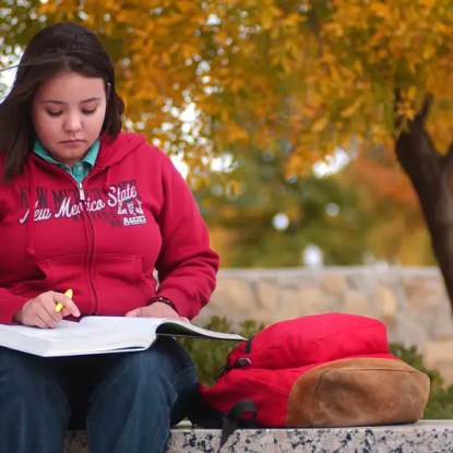 A woman in an NMSU sweatshirt studying on a bench — NMSU Global Campus
