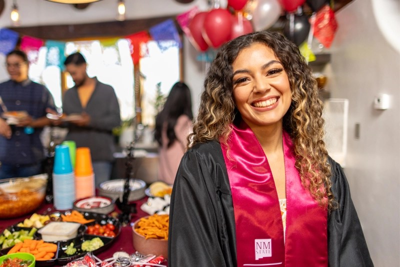 a smiling young woman wearing a graduation stole from New Mexico State University at her graduation party