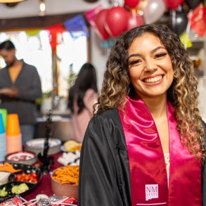 a smiling young woman wearing a graduation stole from New Mexico State University at her graduation party