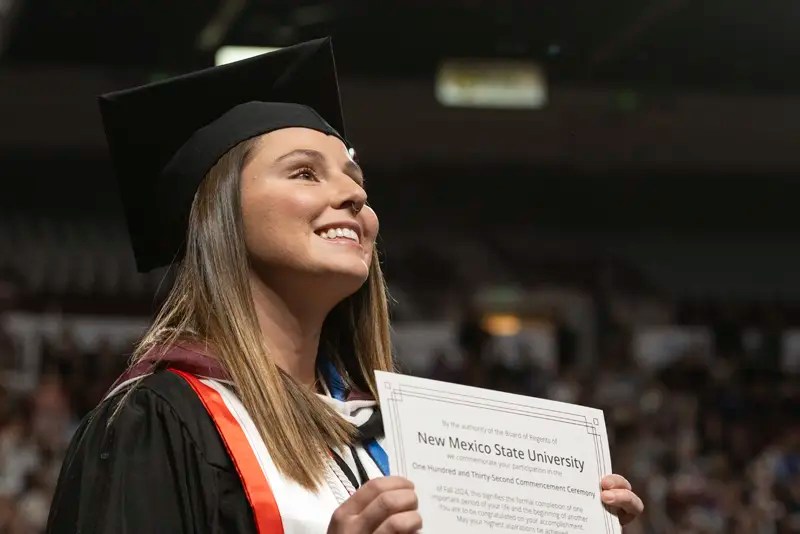 An NMSU graduate in cap and gown holding her diploma — NMSU Global Campus