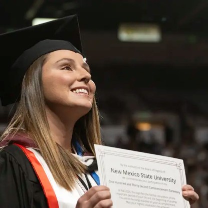 An NMSU graduate in cap and gown holding her diploma — NMSU Global Campus