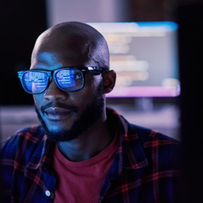 As a man sits in front of a computer, the screen reflects lines of code in his glasses lenses.