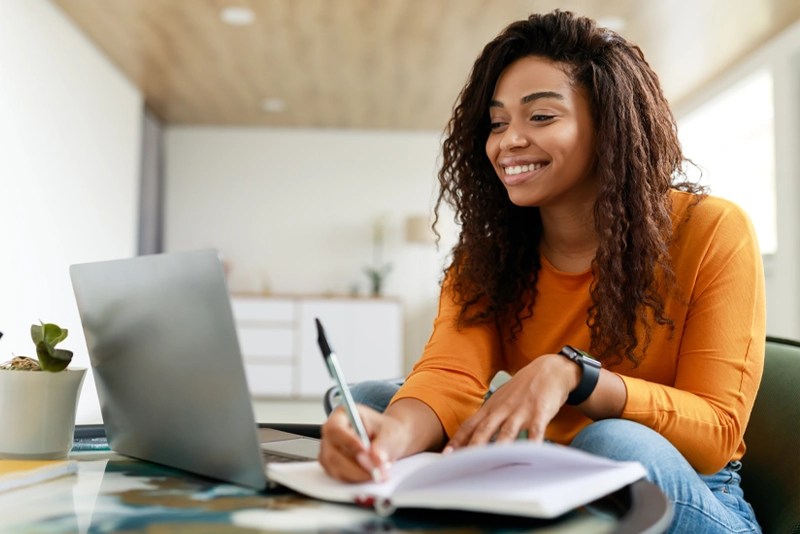 a woman sits at a table using a laptop and a notebook