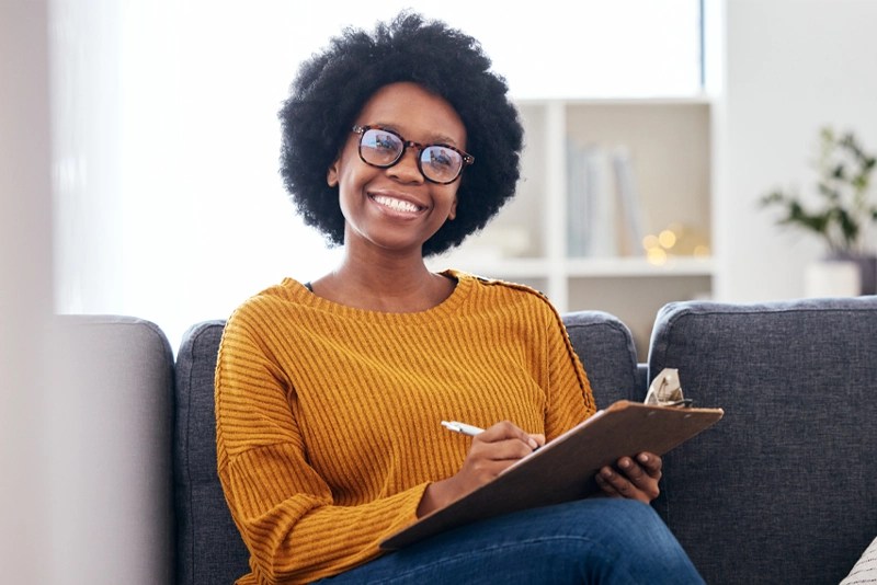 A woman sits on a couch taking notes with a pen and a clipboard