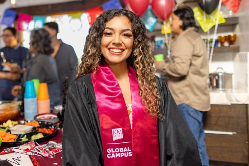 A woman wearing a graduation gown and sash from NMSU Global Campus is smiling at the camera.