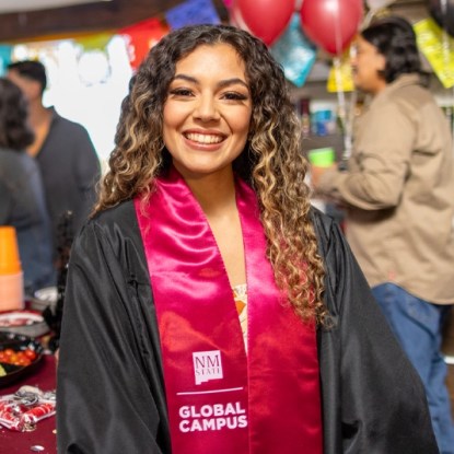 A woman wearing a graduation gown and sash from NMSU Global Campus is smiling at the camera.