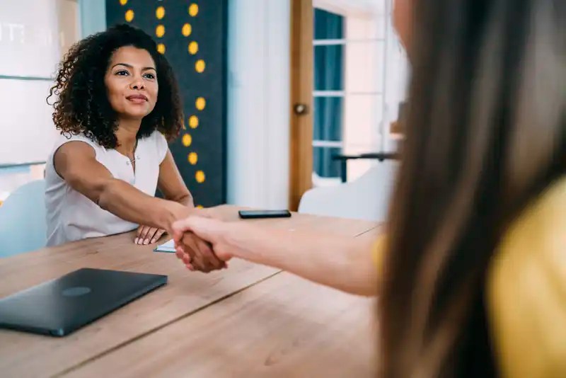 Two women shake hands across a conference room table — New Mexico State University Global Campus