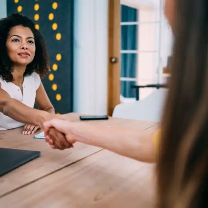 Two women shake hands across a conference room table — New Mexico State University Global Campus