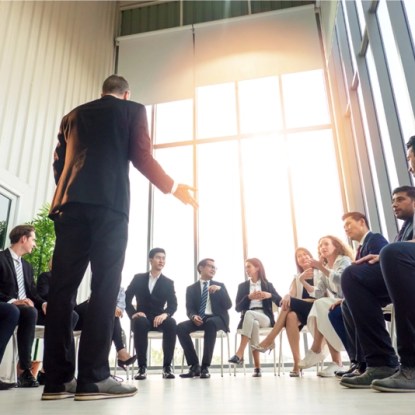 People wearing business suits sit in a circle listening to a man speaking