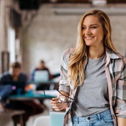 Smiling young woman in a modern coworking space holding eyeglasses, symbolizing confidence, career growth, and learning in a tech startup setting