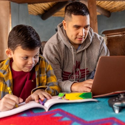 An adult college student works on his laptop while his son colors in a coloring book beside him