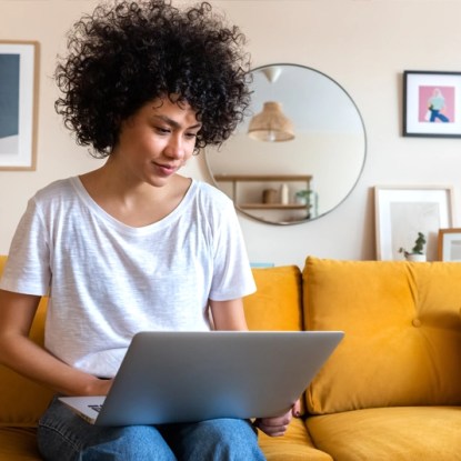 A woman sits on a sofa completing an online application to NMSU Global Campus