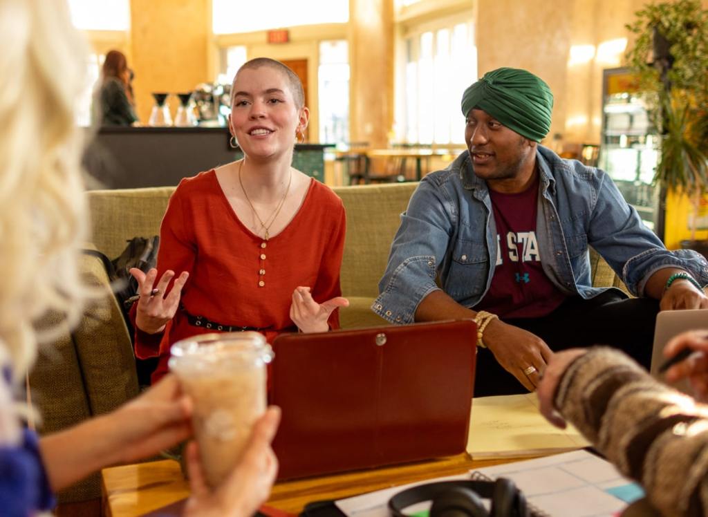 Two people talking in a coffee shop, one with a red blouse and another wearing a green turban