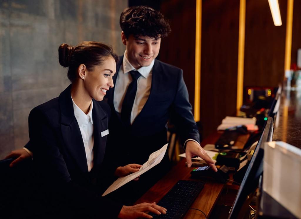Two hotel staff members in uniform smiling and looking at a computer at the front desk