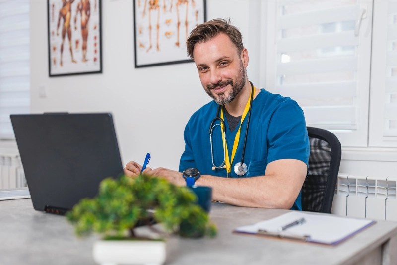 Confident male nurse in scrubs works on a laptop while pursuing an online graduate nursing degree