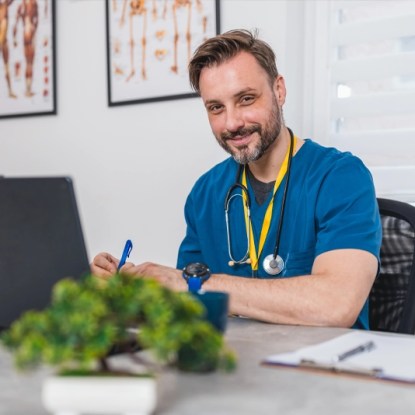 Confident male nurse in scrubs works on a laptop while pursuing an online graduate nursing degree