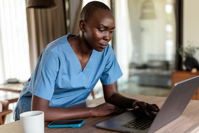 A nurse wearing blue scrubs works on a laptop — NMSU Global Campus