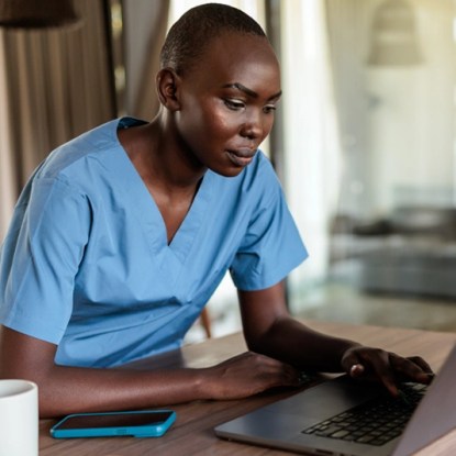 A nurse wearing blue scrubs works on a laptop — NMSU Global Campus