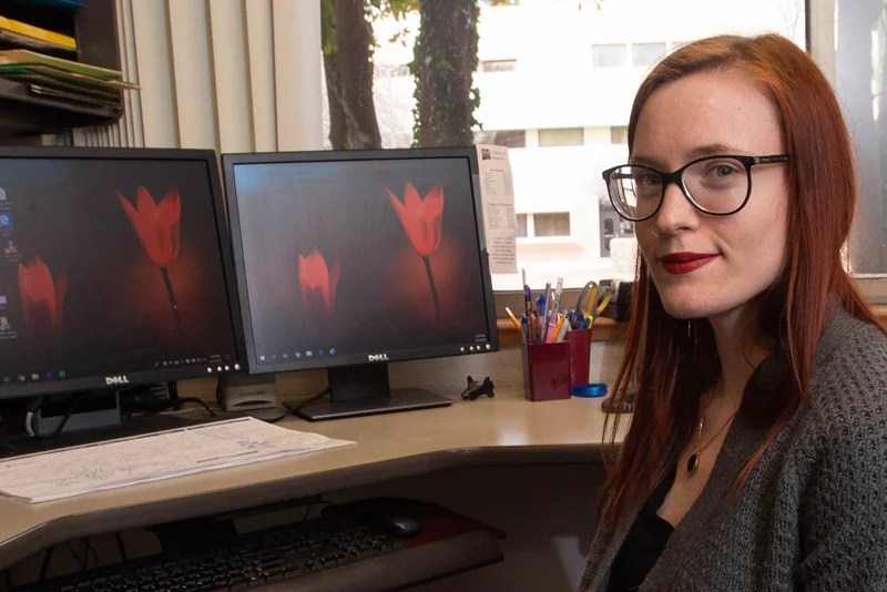 A woman wearing glasses sits at a desk with computer monitors — NMSU Global Campus