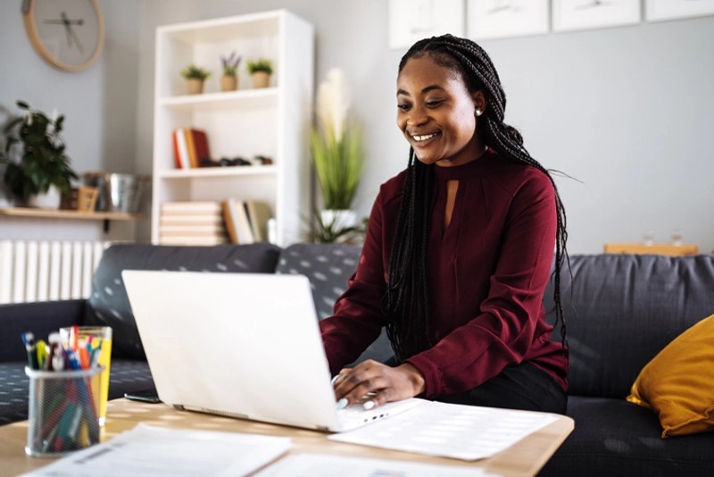 A smiling woman seated on a couch studies comfortably on her laptop — NMSU Global Campus Bachelor of Applied Studies online degree