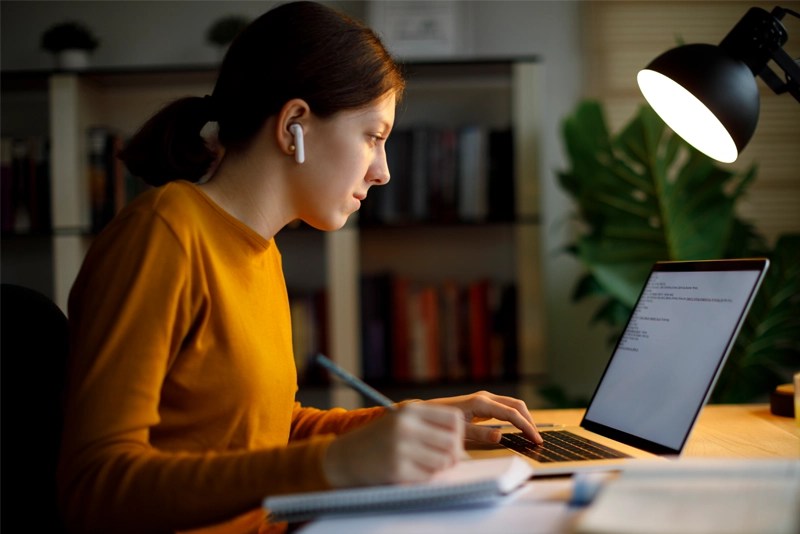 A woman in an orange sweater works on a laptop computer — NMSU Global Campus online communication studies degrees