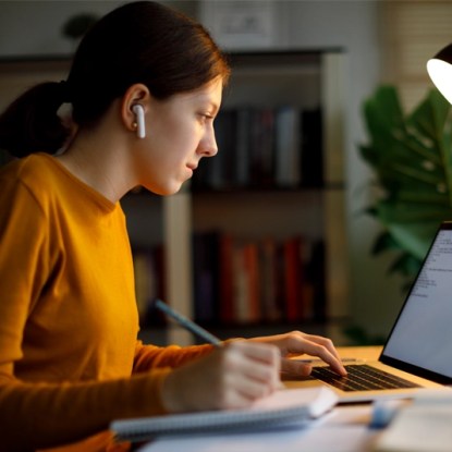 A woman in an orange sweater works on a laptop computer — NMSU Global Campus online communication studies degrees