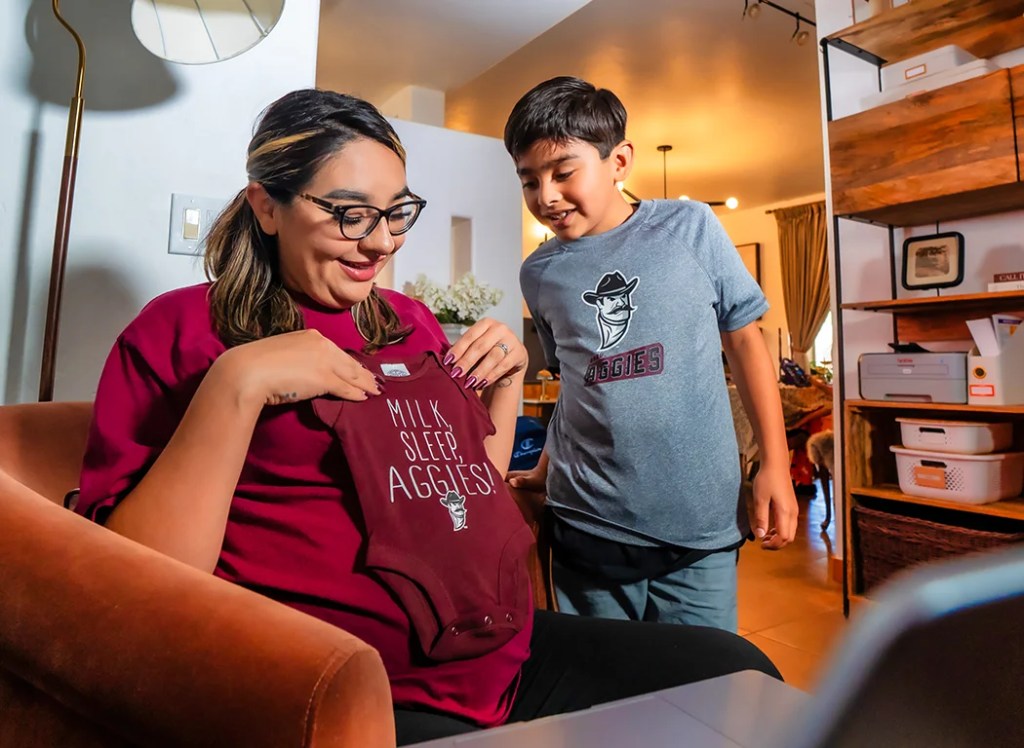 A mother and her young son looking at a crimson NMSU Aggies baby onesie — New Mexico State University Global Campus
