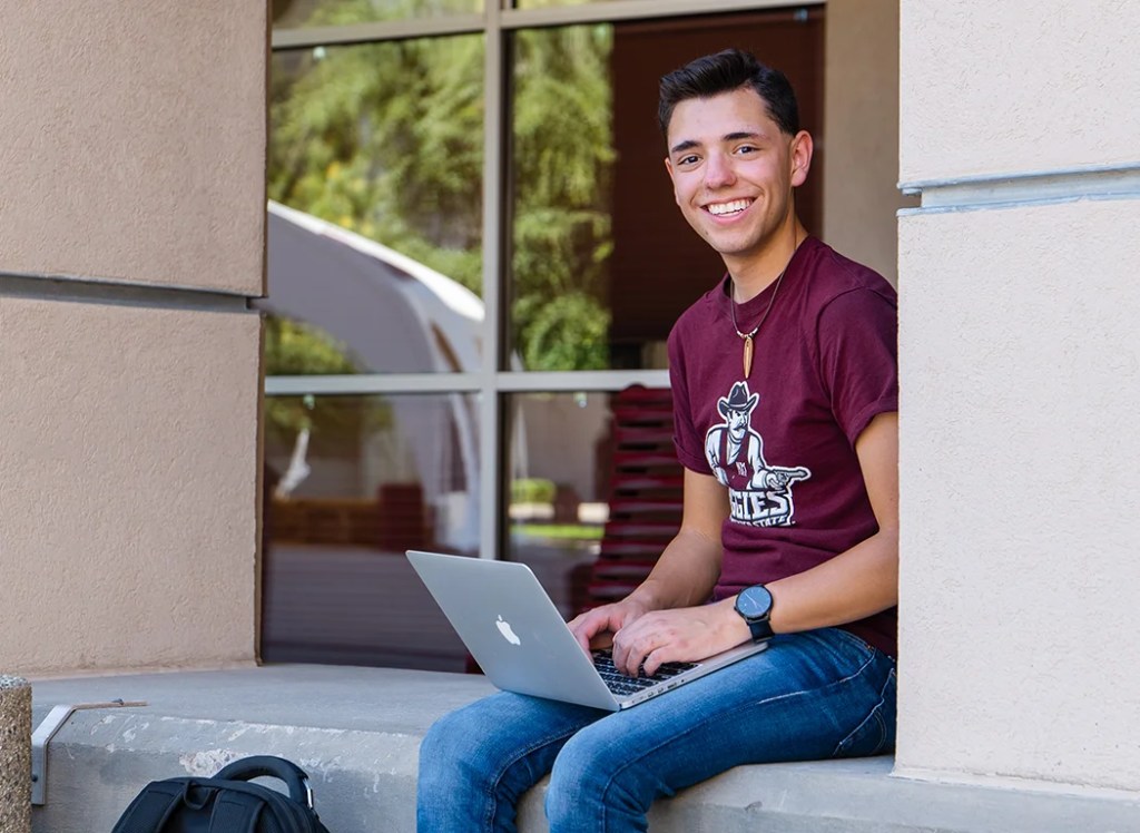 Student in a crimson Aggies t-shirt sits outside studying on his laptop — New Mexico State University Global Campus