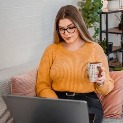 A woman in an orange sweater holds a coffee cup while working on her laptop computer