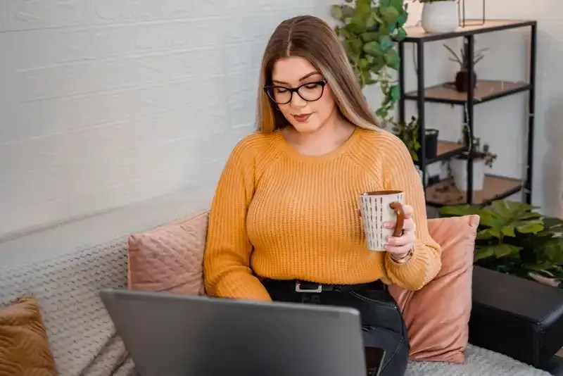 A woman in an orange sweater holds a coffee cup while working on her laptop computer