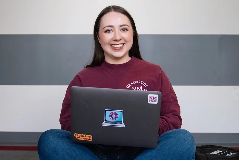 A smiling NMSU Global Campus student sits crosslegged with her laptop