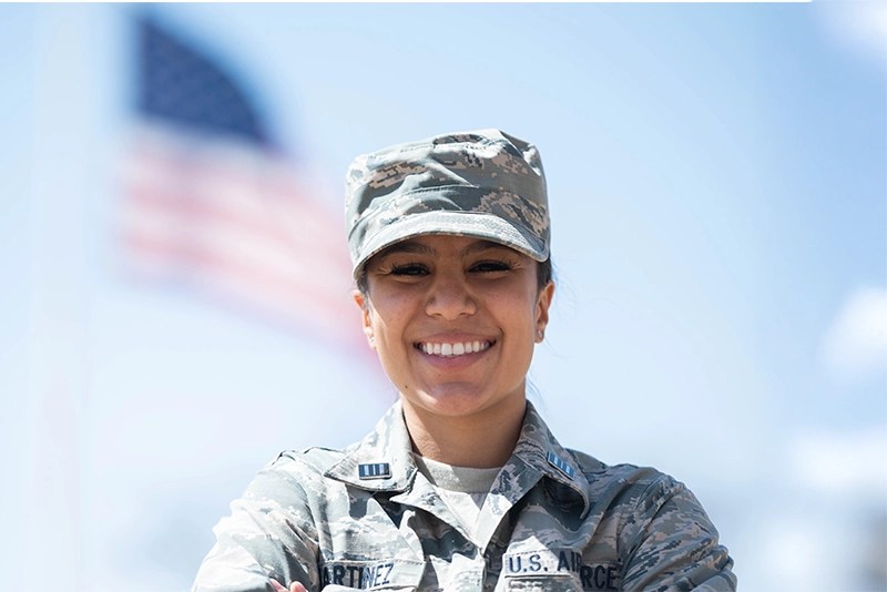 A smiling woman in military fatigues with the U.S. flag waving in the background