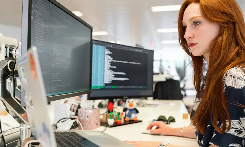 A woman working at a desk with two large computer monitors