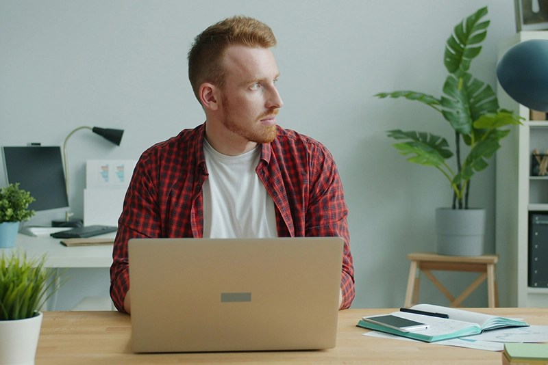 A man looking out a window pensively as he types on his laptop