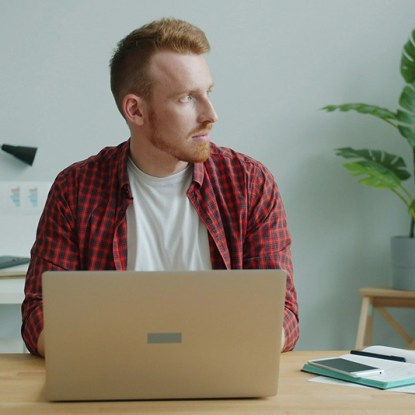 A man looking out a window pensively as he types on his laptop