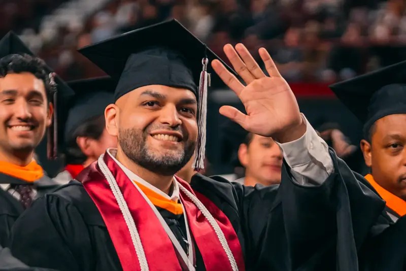 A man in graduation regalia waving amongst a crowd of other graduates