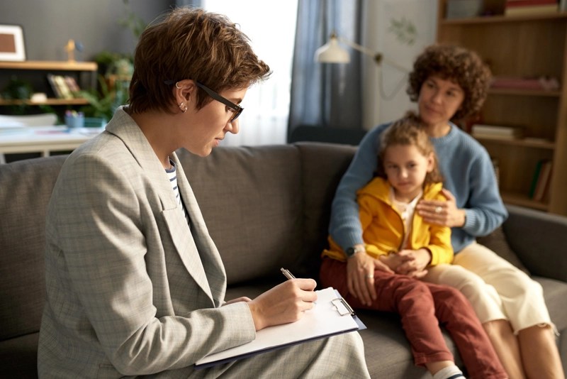 A school social worker taking notes while talking with a mother and her young daughter on a sofa
