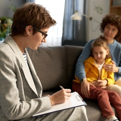 A school social worker taking notes while talking with a mother and her young daughter on a sofa