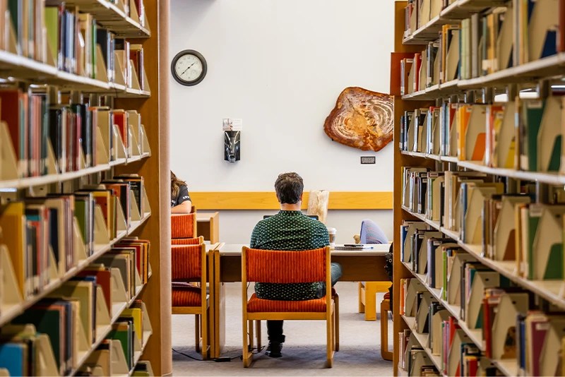 College students studying at a table between bookshelves in a university library.